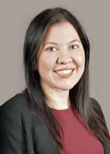 A woman with long dark hair, wearing a maroon top and black blazer, smiles at the camera against a plain gray background.