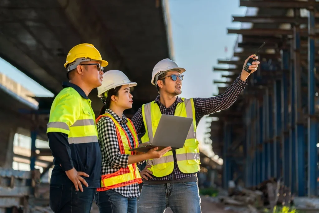 Three construction workers in safety gear and hard hats stand under a bridge; one holds a laptop, another gestures with a walkie-talkie, and they appear to be discussing the project.