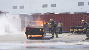 Firefighters spray water on a burning vehicle during a training exercise in an outdoor facility, with observers and equipment in the background.