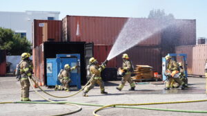 Firefighters in protective gear spray water from a hose during a training exercise near shipping containers and industrial equipment outdoors.