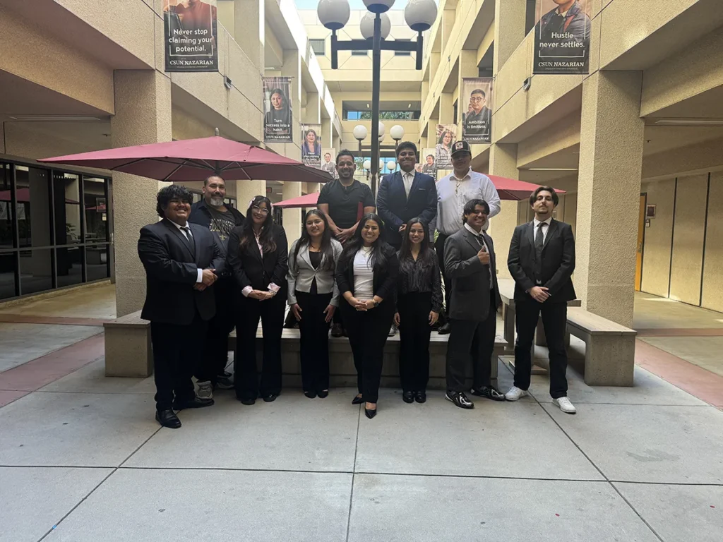 A group of twelve people in business attire pose for a photo in an outdoor courtyard with umbrellas and posters hanging above.