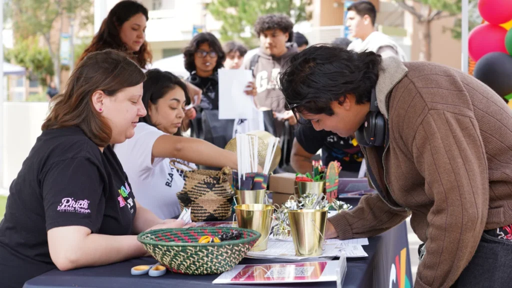 People standing and sitting at an outdoor event table, filling out forms and picking up materials, with baskets, gold buckets, and decorations on the table.