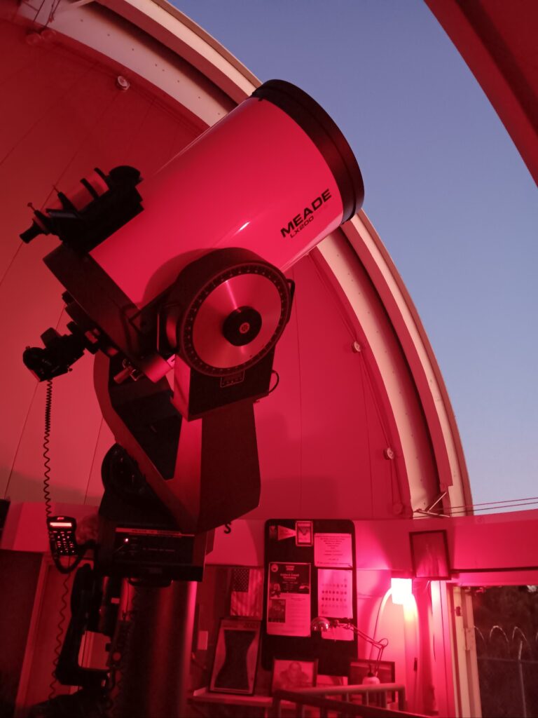 Large Meade telescope inside an observatory dome at dusk, illuminated by red lights, with informational posters visible in the background.