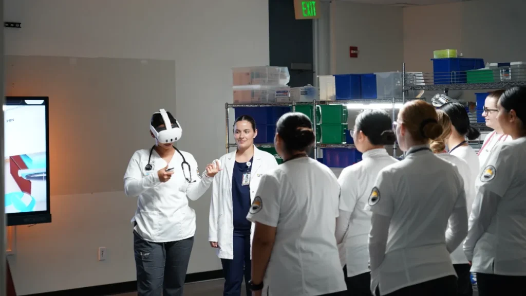 A group of medical students watch as one person uses a virtual reality headset in a classroom setting, with an instructor standing nearby.