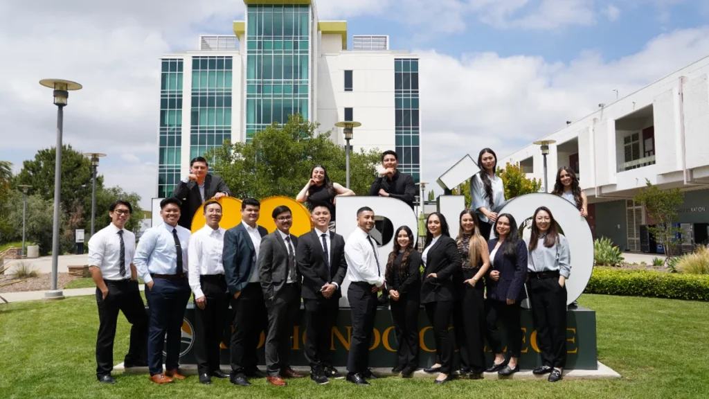 A group of professionally dressed young adults pose for a photo in front of a college campus building and a large sculpture outdoors.
