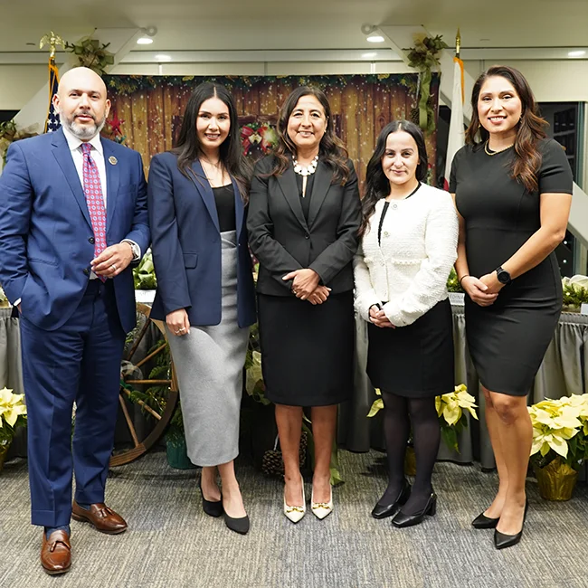 Five people dressed in business attire stand indoors in front of a decorated table with plants and flags visible in the background.