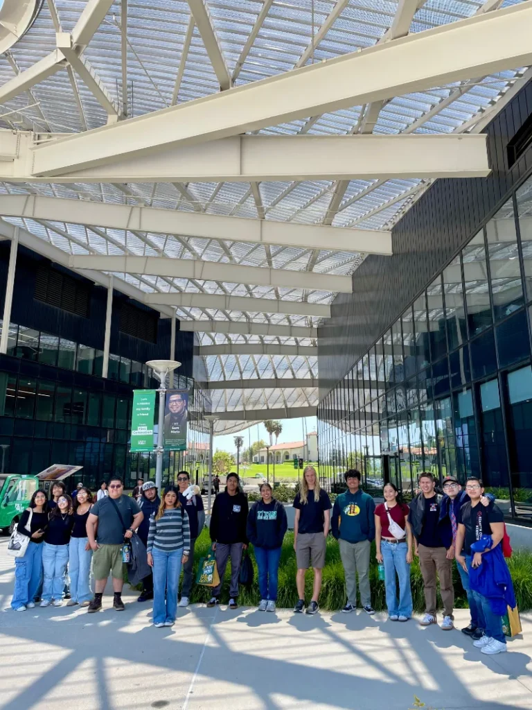 A group of people poses for a photo outside a modern building with glass walls and a large metal canopy on a sunny day.