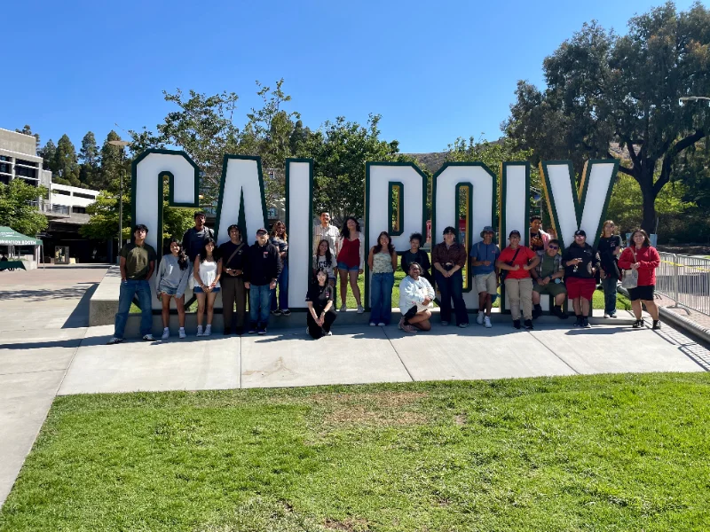 A group of people pose in front of large "Cal Poly" letters on a concrete walkway, with trees and campus buildings in the background.