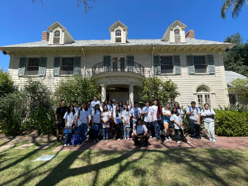 A group of people pose for a photo on the lawn in front of a large, light-colored house with a balcony and green shutters on a sunny day.