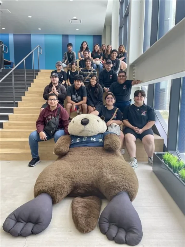 A group of people poses on stairs behind a large otter mascot plush wearing a "SUMA" shirt, in a bright indoor space with large windows.