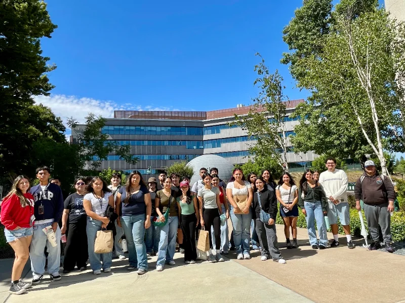 A group of people stands in a line outside on a sunny day, with modern buildings and trees in the background.