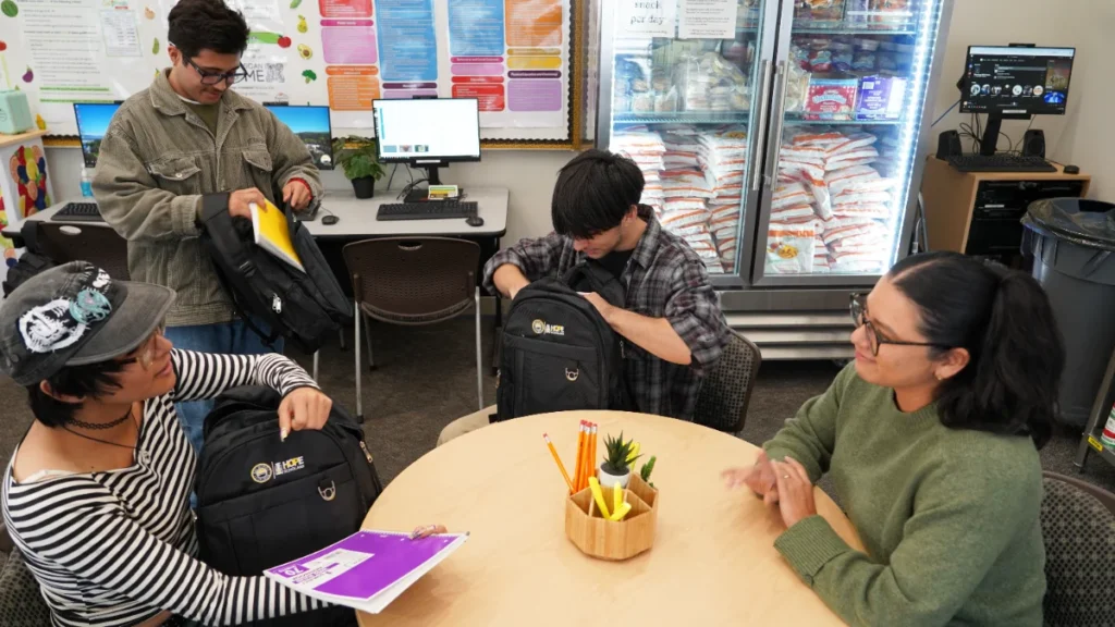 Four students sit around a table; two unpack backpacks, one holds notebooks, and another sits with hands on the table. Bags of rice are visible in a fridge behind them.