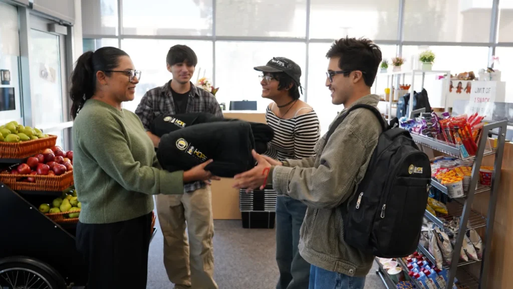 Four people stand in a store near shelves of snacks and apples; one person hands a rolled blanket with a logo to another person with a backpack.
