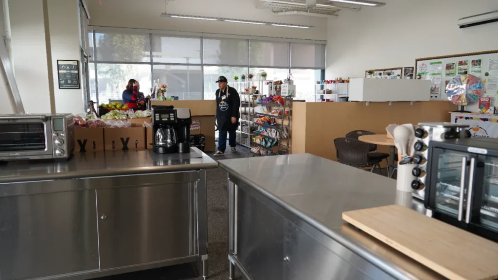 Two people work in a small grocery or café with stainless steel counters, a coffee maker, shelves of food, and boxed produce near large windows.