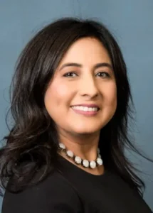 A woman with medium-length dark hair, wearing a black top and a beaded necklace, smiles at the camera against a plain blue background.