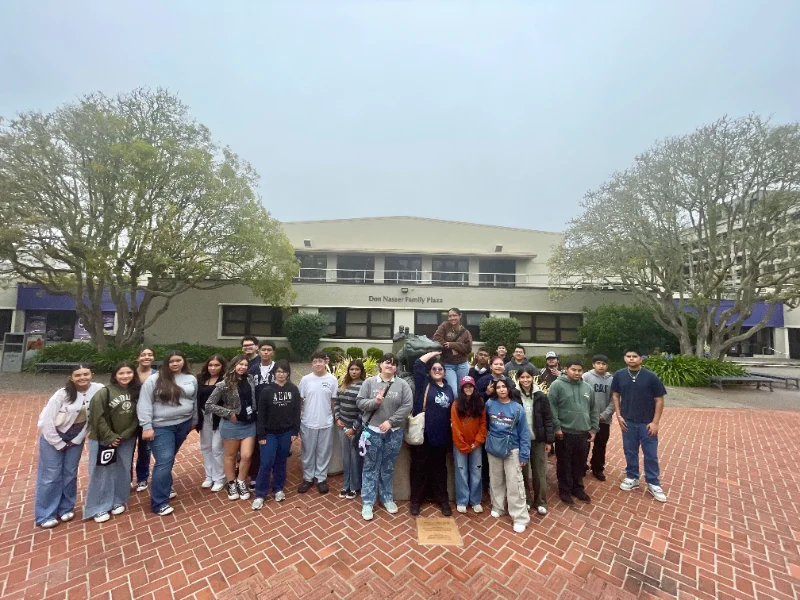 A group of people pose for a photo in front of a two-story building with the sign "Don Nasser Family Plaza" on a foggy day.