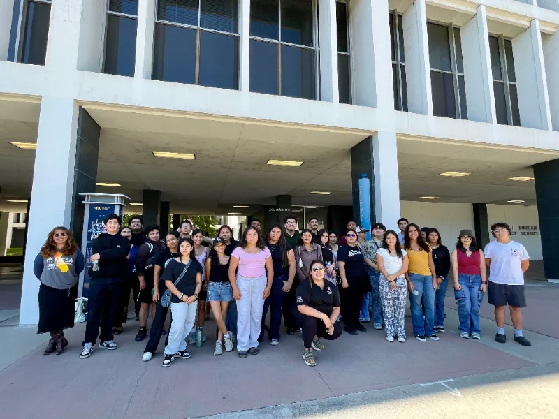 A group of students poses for a photo in front of a building with large windows and columns on a sunny day.