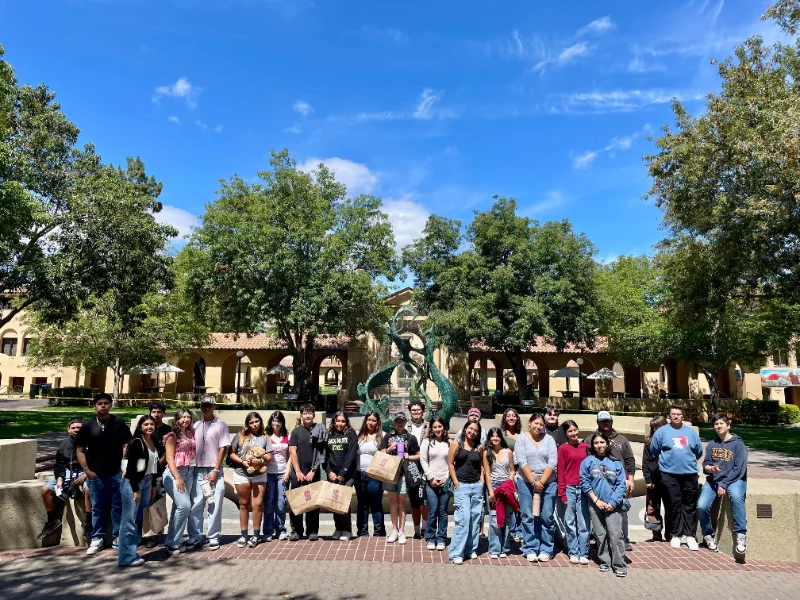 A group of people stands in a line outdoors in front of a serpentine statue, holding bags and posing for a group photo on a sunny day with trees and buildings in the background.