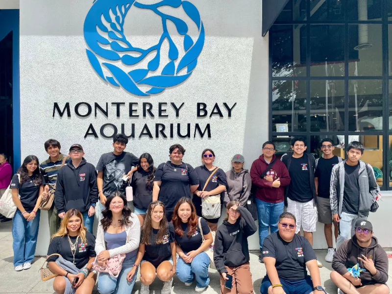 A group of people poses for a photo in front of the Monterey Bay Aquarium entrance sign on a sunny day.