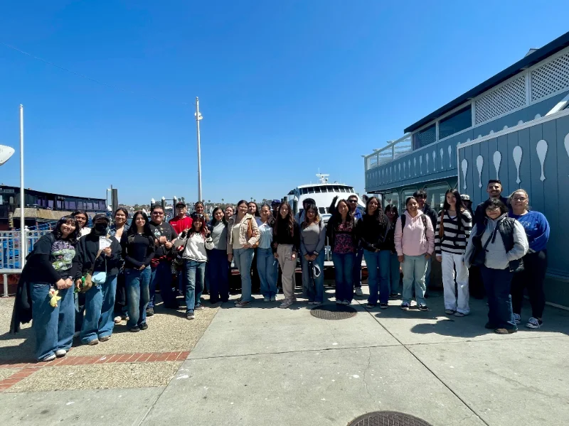 A group of people stands together outdoors on a sunny day in front of a boat, with a building and blue sky in the background.