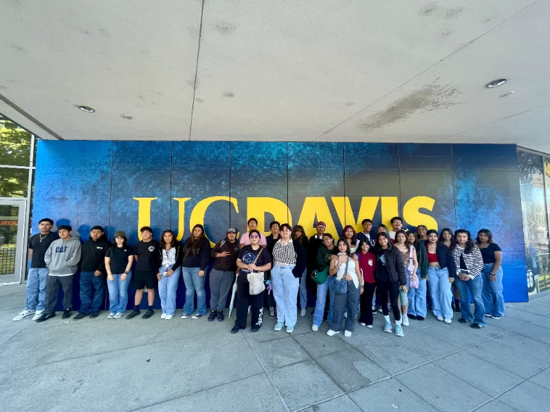 A group of students poses for a photo in front of a large UC Davis sign on a blue and yellow background.