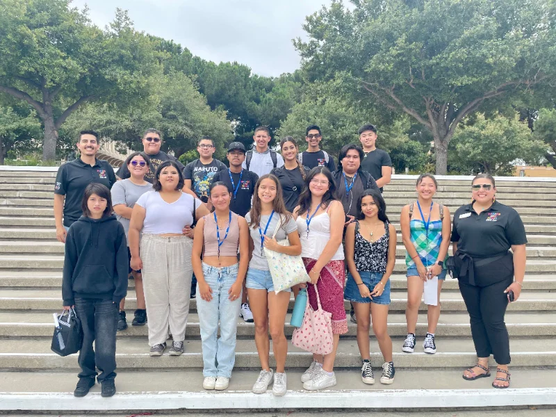 A group of young people and two adults stand on outdoor steps, posing for a photo with trees in the background.