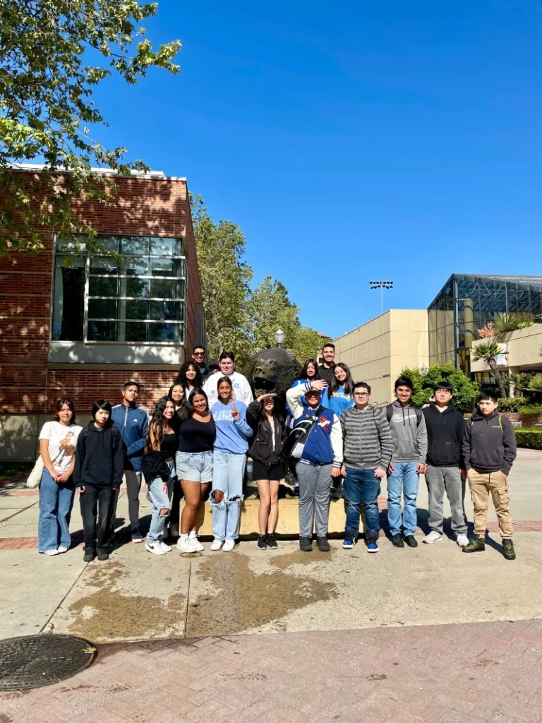 A group of 18 people stands in front of a bronze bear statue on a sunny day, with campus buildings and trees visible in the background.