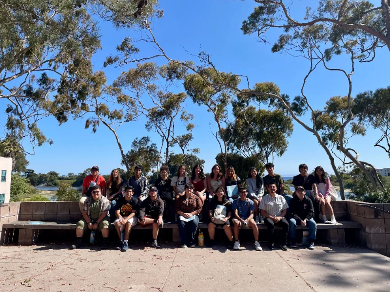 A group of people sit on a long stone bench outdoors in front of tall trees and a clear blue sky.