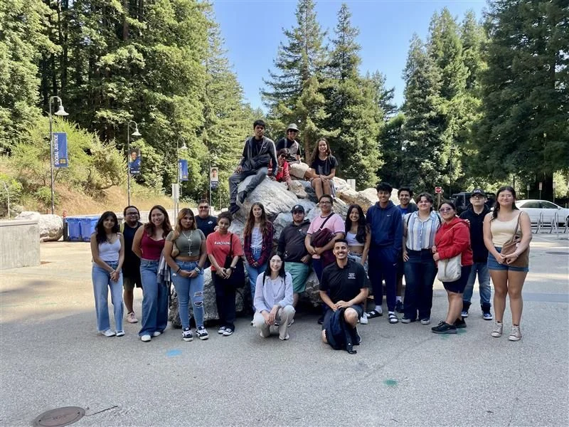 A group of people pose together in front of a large rock outdoors, surrounded by trees and blue sky.