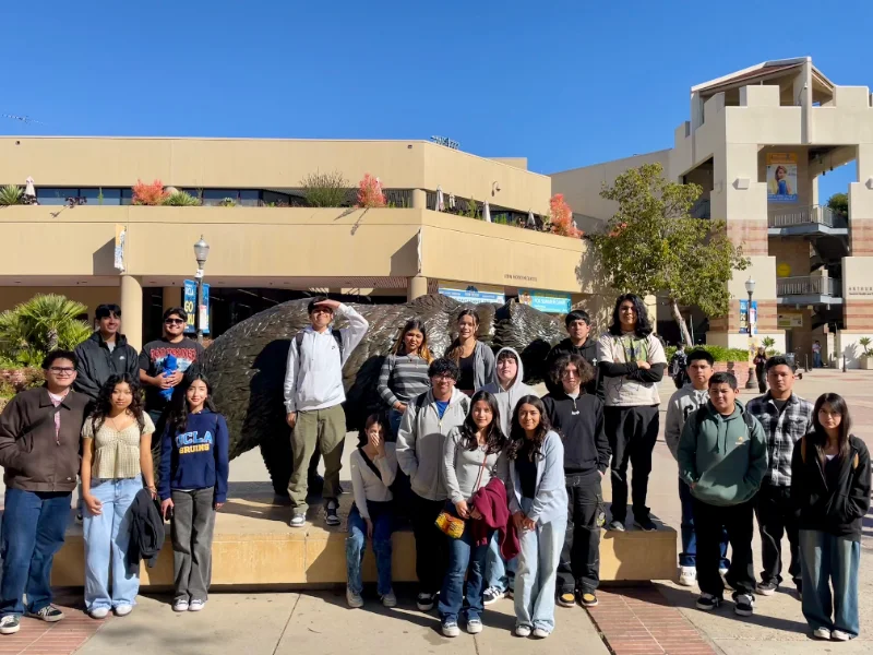 A group of young adults poses in front of a bear statue on a sunny day, with campus buildings and trees in the background.