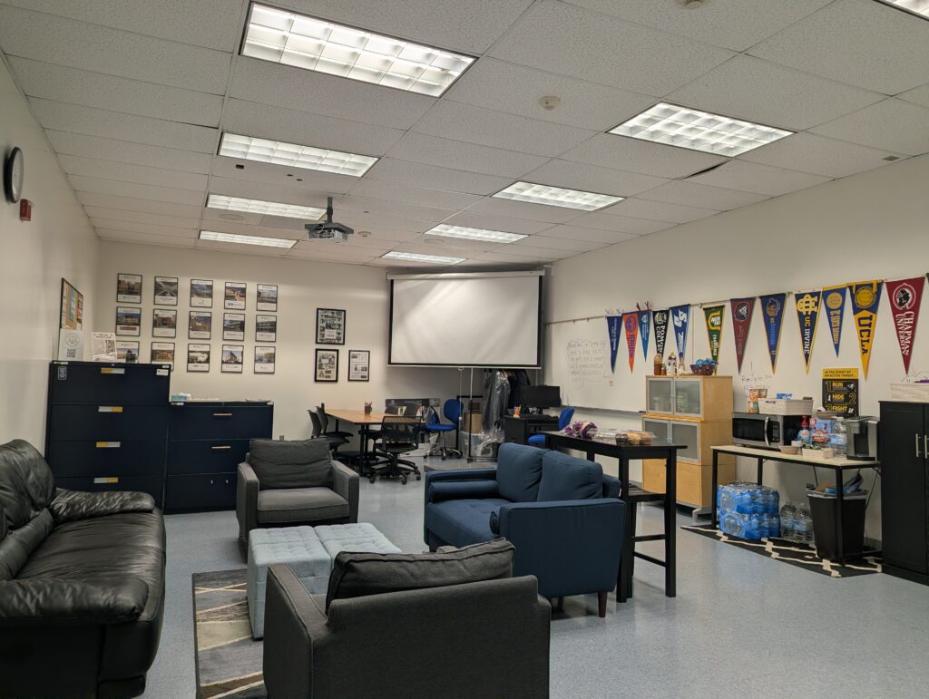 A classroom or lounge with sofas, armchairs, a projector screen, framed photos, college pennants on the wall, and a water cooler in the corner.
