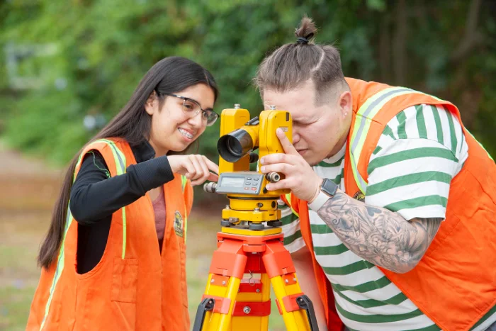 Two people in orange safety vests use a yellow surveying instrument outdoors, with one looking through the viewfinder and the other adjusting the device.