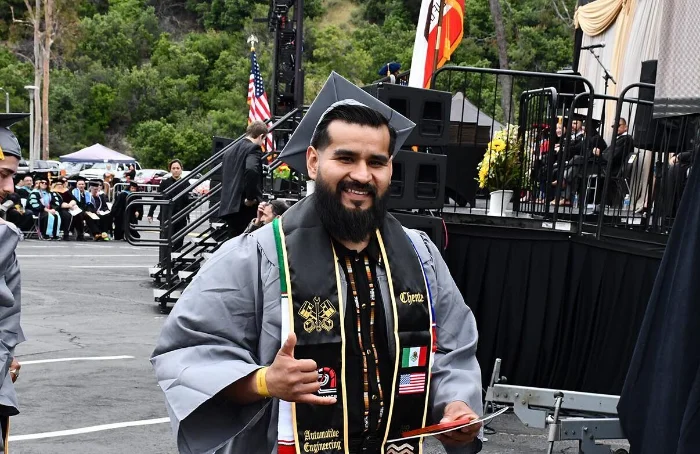 A graduate in a cap and gown smiles and gives a thumbs up while holding his diploma at an outdoor graduation ceremony.
