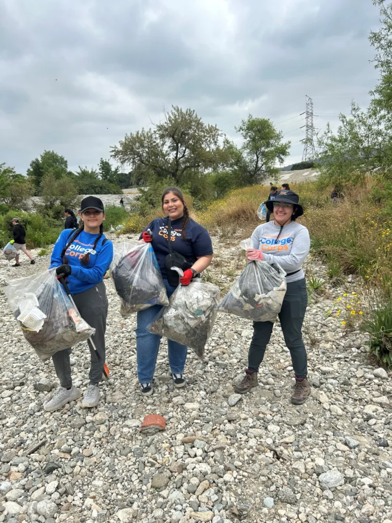 Three people stand outdoors on a rocky riverbed, each holding a large bag of collected trash, participating in a cleanup event.
