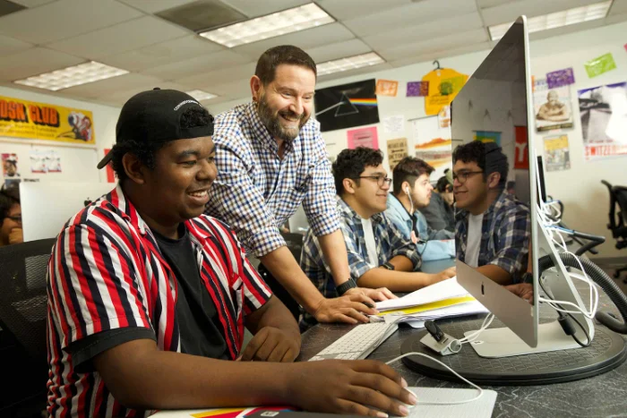A teacher stands beside students working on computers in a classroom, smiling as he assists them with their assignments.