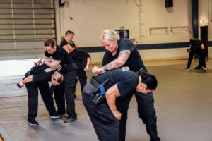 A group of people in uniform practice self-defense techniques in a gymnasium, with pairs engaged in physical maneuvers on padded flooring.