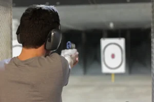 A person wearing ear protection aims a handgun at a paper target at an indoor shooting range.