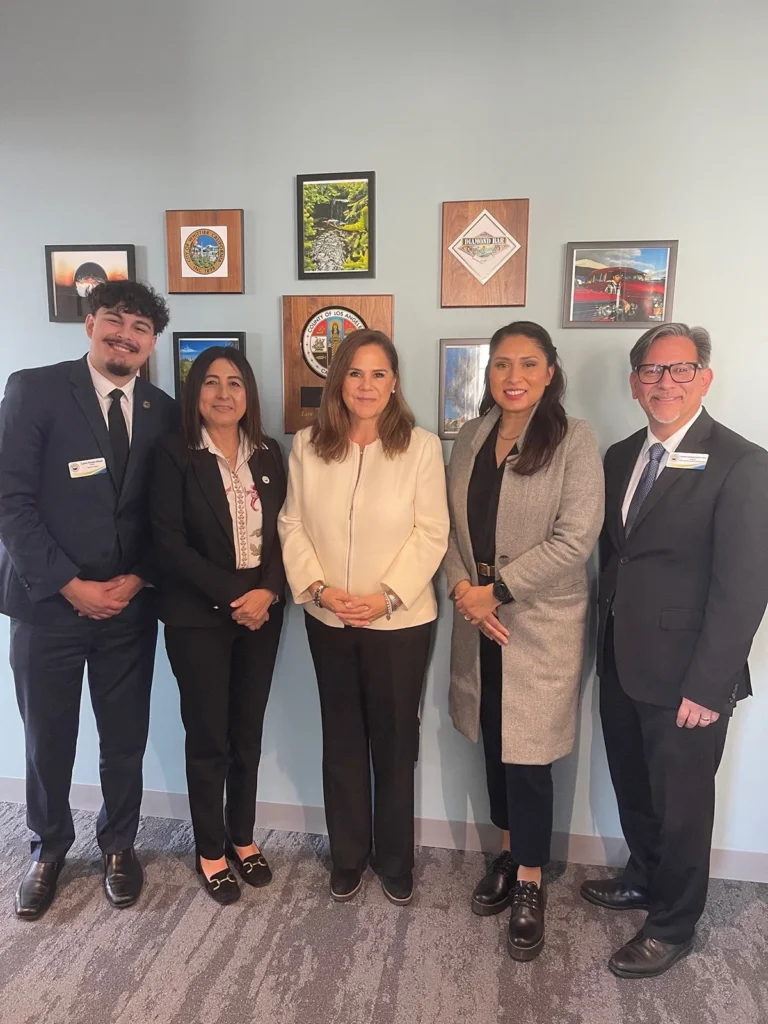 Five people stand in a row, dressed in business attire, in front of a wall decorated with framed pictures and logos.