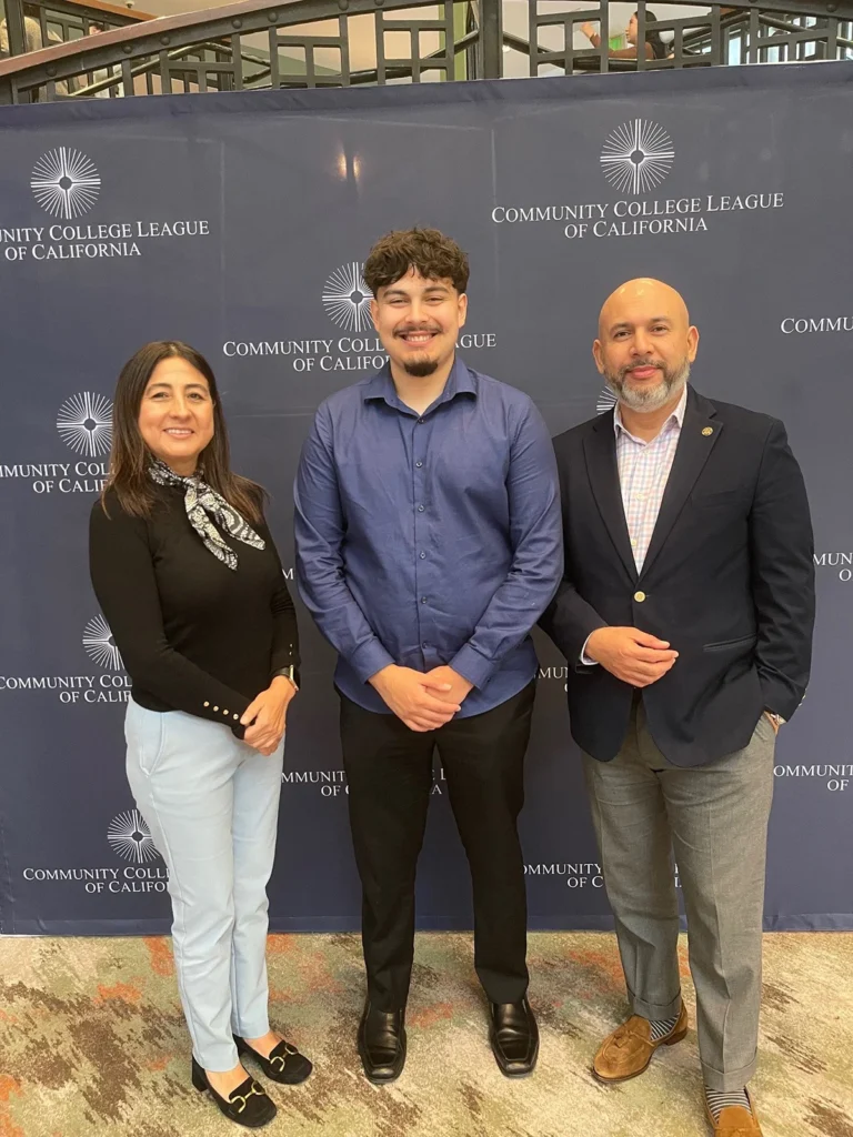 Three people standing and facing the camera in front of a "Community College League of California" backdrop.