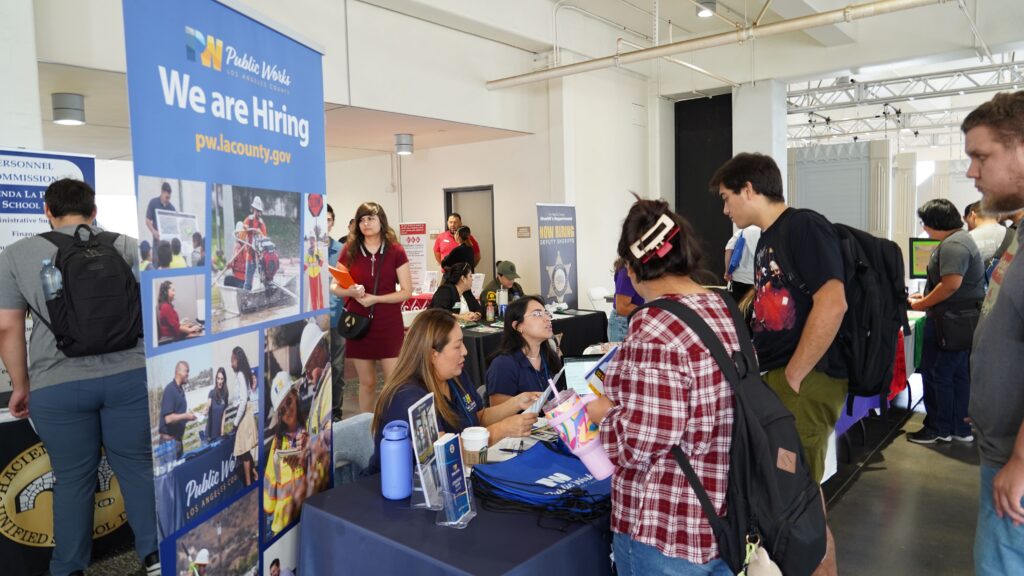 People interact at a job fair. A "We are Hiring" sign for Public Works is visible, with tables, informational materials, and attendees talking with recruiters.
