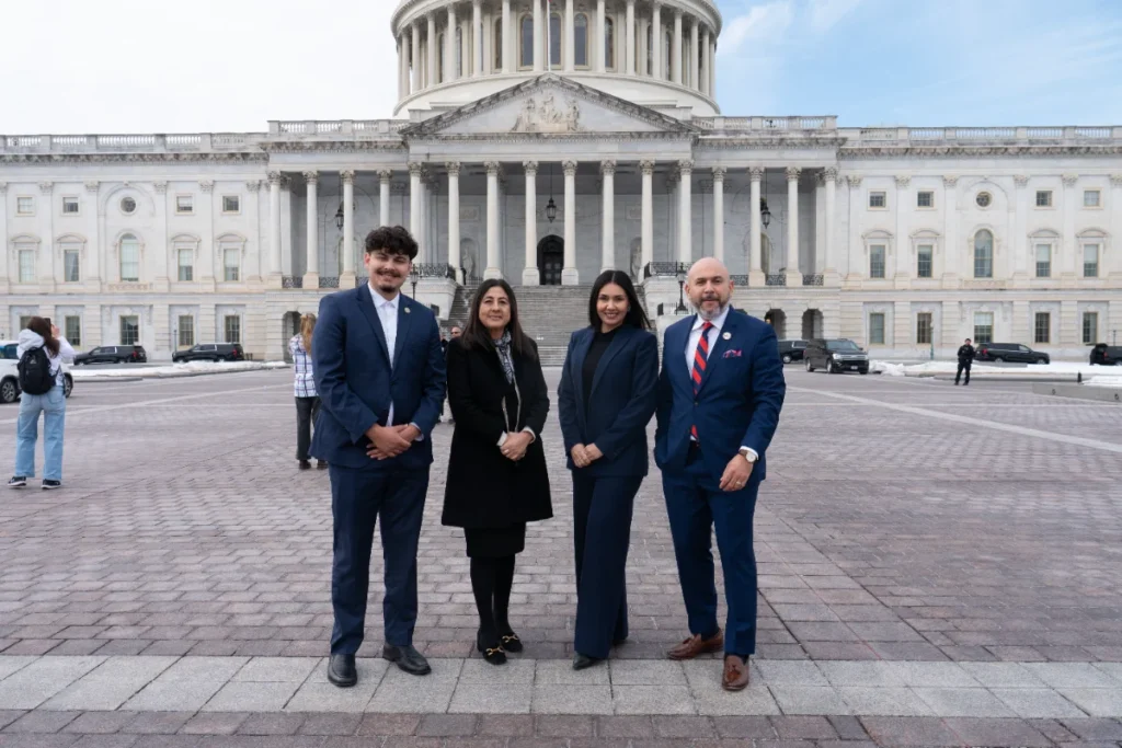 Four people in business attire stand in front of the U.S. Capitol building on a paved plaza, posing for a group photo.