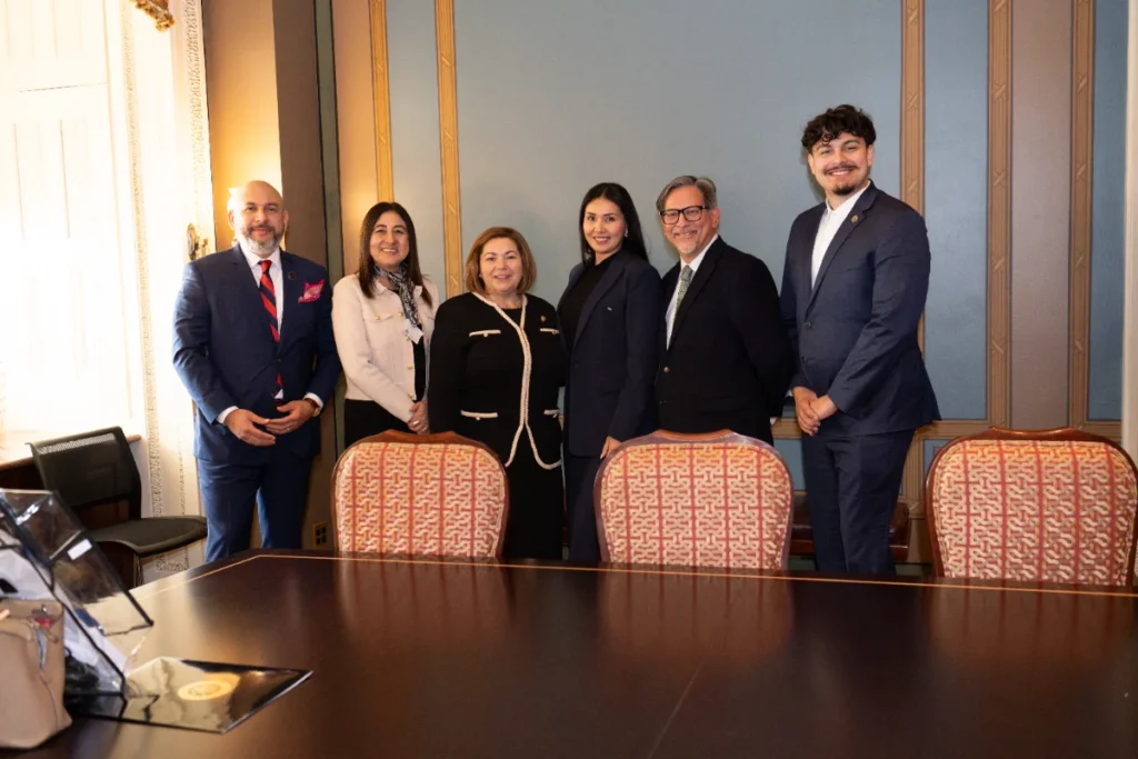 Six people in business attire stand together in a formal meeting room, smiling at the camera. A large wooden table with patterned chairs is in the foreground.