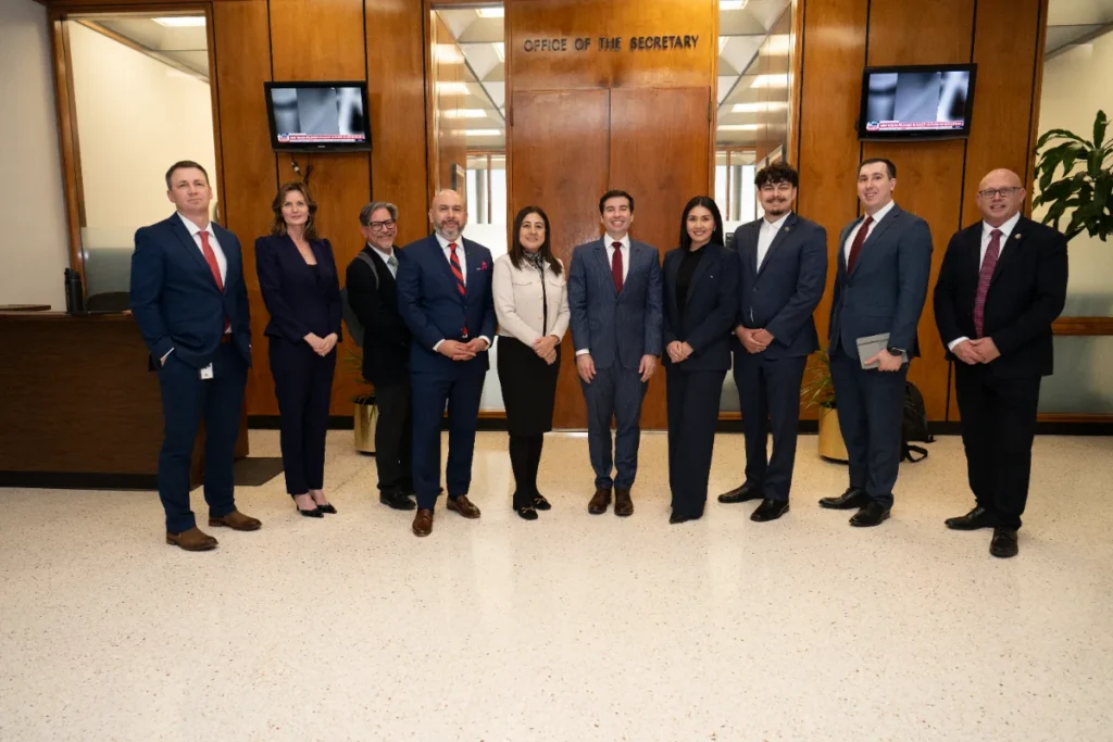 Ten people in business attire stand in a row, posing for a group photo inside an office lobby labeled "Office of the Secretary.