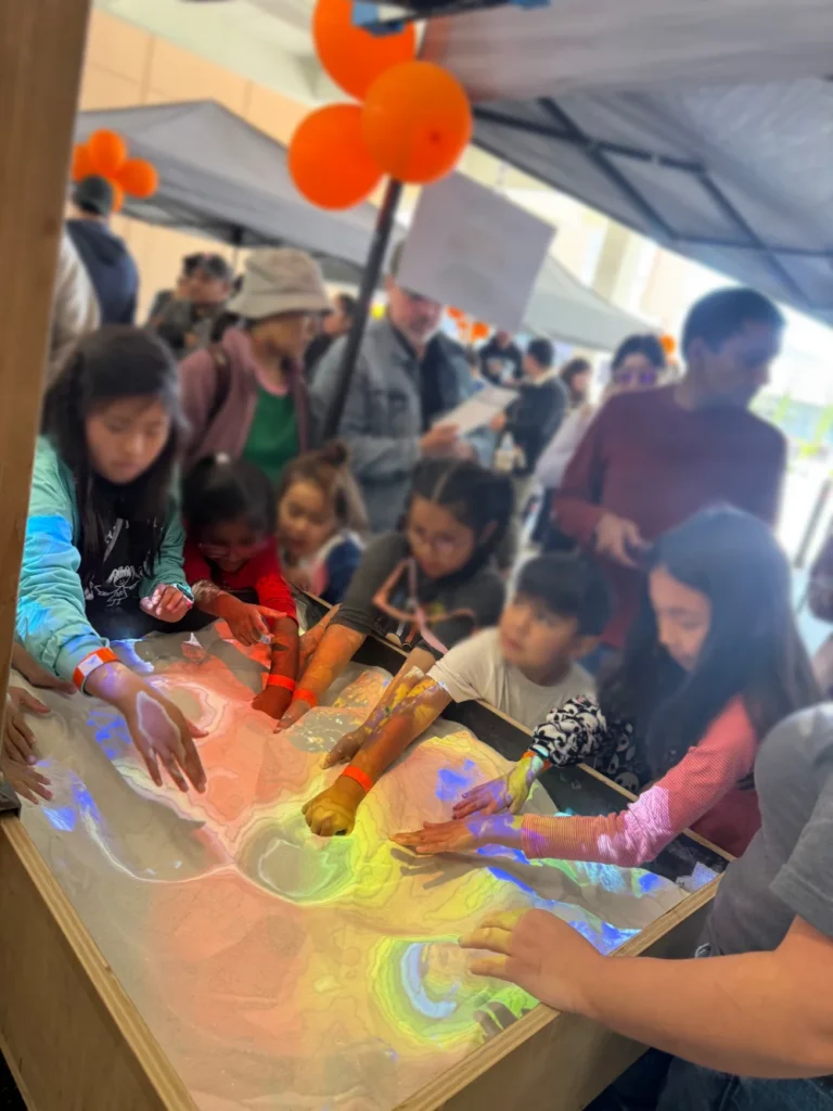 Children interact with a digital sandbox exhibit, shaping sand as projected colors display topographic patterns at a busy indoor event.