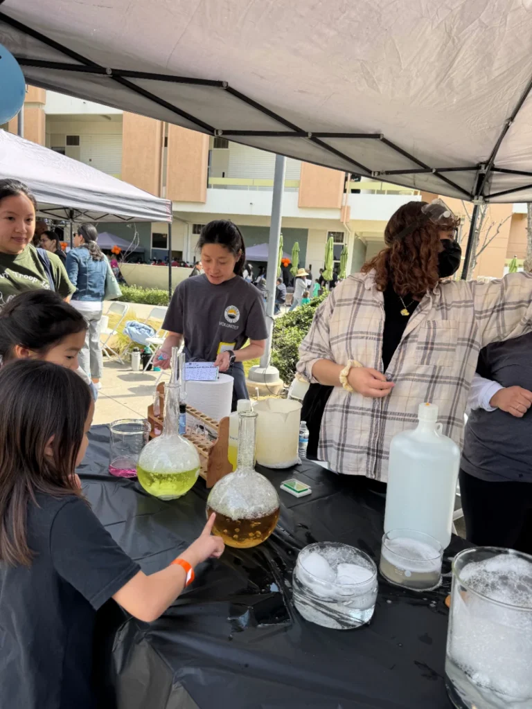 Children and adults gather at a booth with science equipment, including flasks with colored liquids and dry ice, under a canopy at an outdoor event.