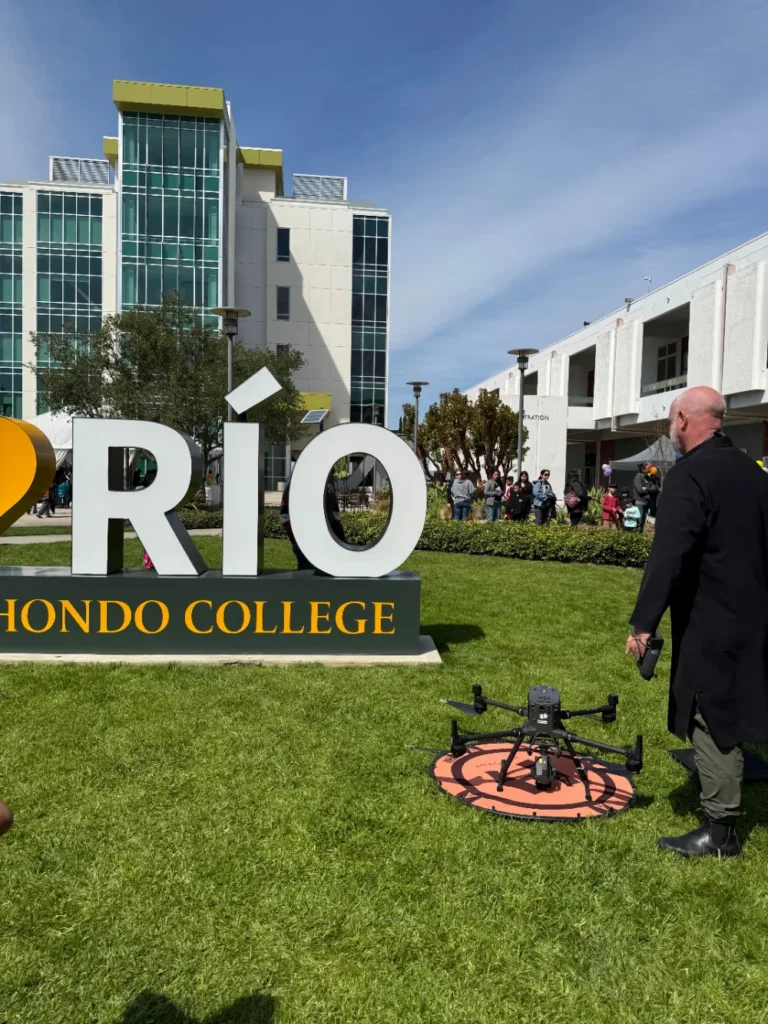 A person in a black coat stands near a drone on a landing pad in front of a large Rio Hondo College sign, with a modern building and crowd in the background.
