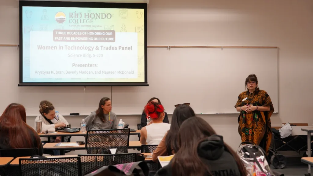 A presenter speaks to an audience during the "Women in Technology & Trades Panel" at Rio Hondo College; the event title and presenters' names are displayed on a projector screen.