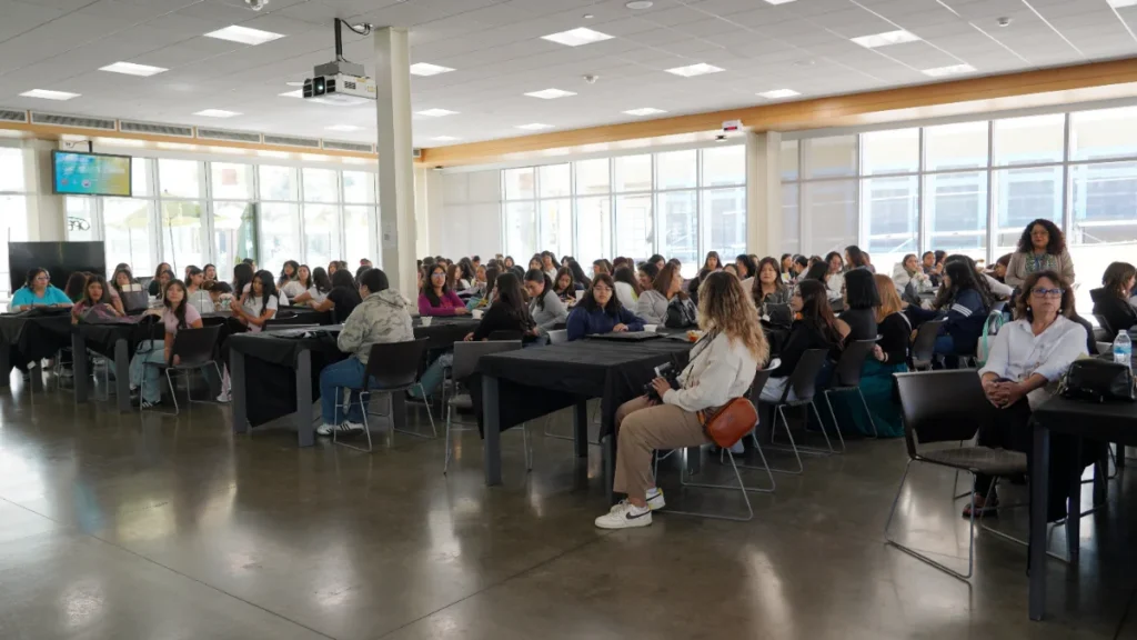 A large group of people sit at tables in a bright, modern room with floor-to-ceiling windows during what appears to be a meeting or event.
