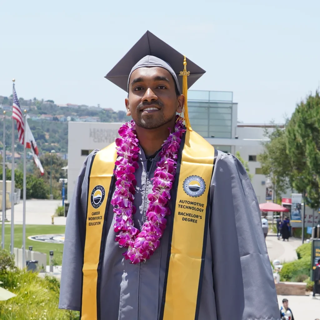 A graduate wearing a gray cap and gown, yellow stoles, and a purple lei stands outdoors on a campus with flags and buildings in the background.