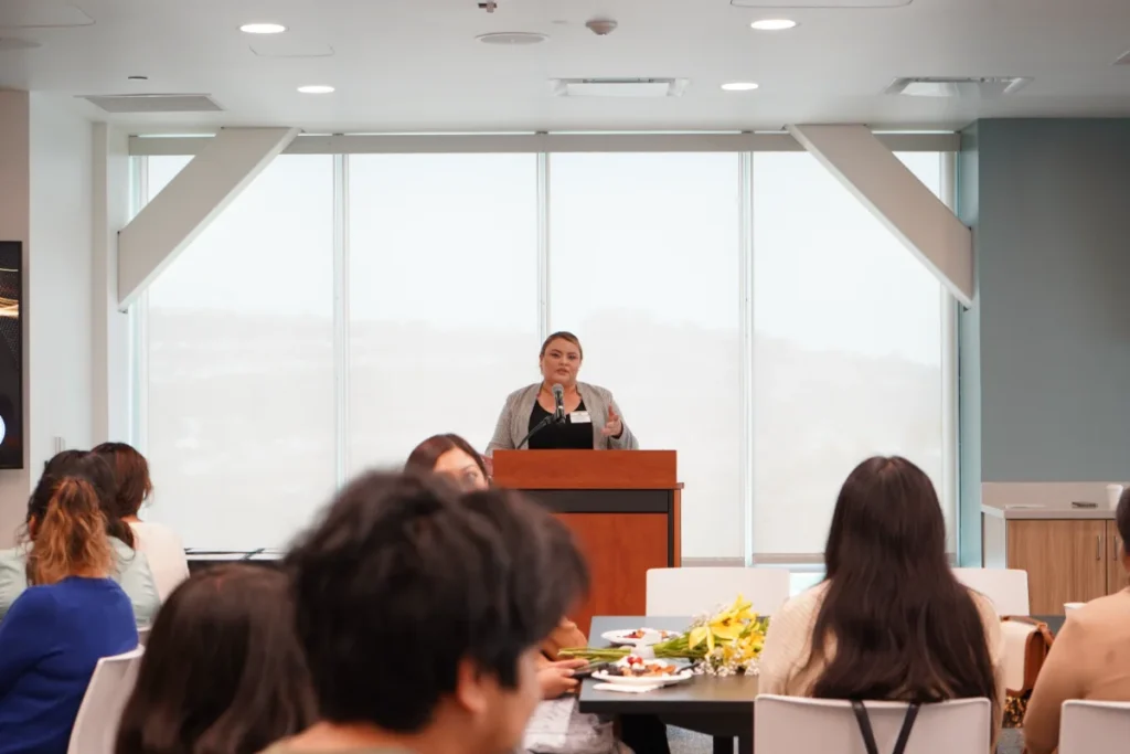 A woman stands at a podium speaking to an audience seated at tables in a brightly lit room with large windows.